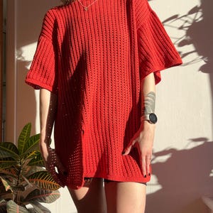 May include: A bright red, oversized crochet top with short sleeves. The top has a loose, open weave design, creating a textured appearance. The garment is knee-length and appears to be handmade. The top is a vibrant, solid color.