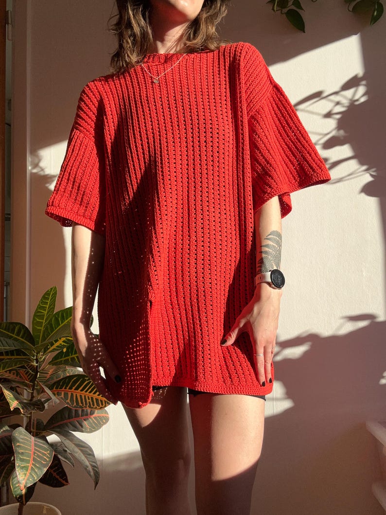 May include: A bright red, oversized crochet top with short sleeves. The top has a loose, open weave design, creating a textured appearance. The garment is knee-length and appears to be handmade. The top is a vibrant, solid color.