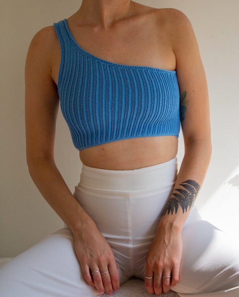 May include: A light blue one-shoulder crop top made of a ribbed knit fabric. The top is worn with white trousers.
