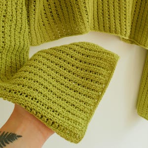 May include: A green crochet sweater with a loose, open knit pattern. The sweater has long sleeves and a cropped length.