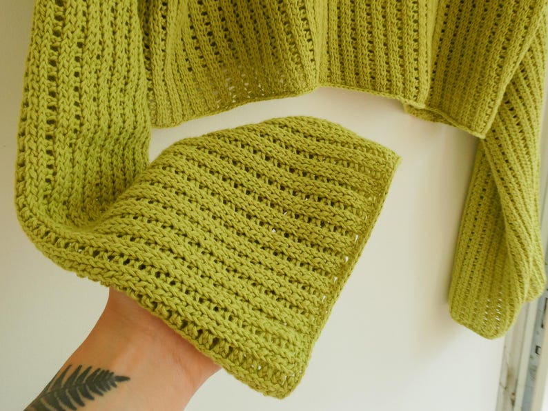 May include: A green crochet sweater with a loose, open knit pattern. The sweater has long sleeves and a cropped length.