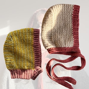 May include: Two handmade crochet bonnets. One bonnet is cream and burgundy, with long ties. The other bonnet is yellow and green striped with a burgundy trim. Both bonnets are made of a textured yarn.