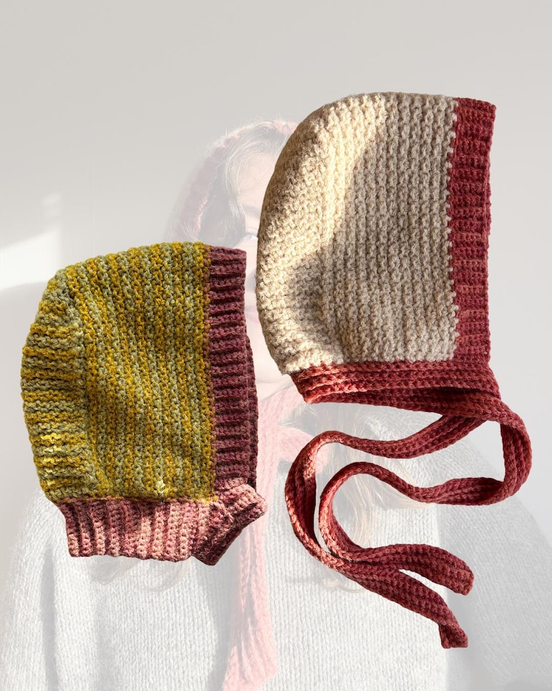 May include: Two handmade crochet bonnets. One bonnet is cream and burgundy, with long ties. The other bonnet is yellow and green striped with a burgundy trim. Both bonnets are made of a textured yarn.