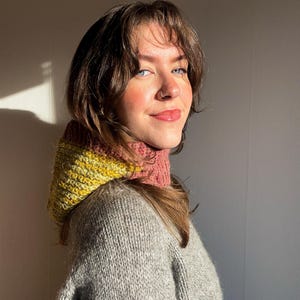 May include: A cozy, hand-knitted cowl in shades of yellow, pink, and gray. The cowl is worn over a gray sweater, creating a warm and stylish look. The knitted texture is visible, highlighting the craftsmanship of the piece. The overall aesthetic is soft and inviting.