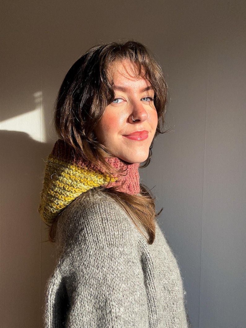 May include: A cozy, hand-knitted cowl in shades of yellow, pink, and gray. The cowl is worn over a gray sweater, creating a warm and stylish look. The knitted texture is visible, highlighting the craftsmanship of the piece. The overall aesthetic is soft and inviting.