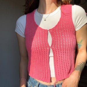 May include: A bright pink, open-front knit vest with a delicate, open weave. The vest is worn over a white t-shirt and paired with blue jeans. The vest features a drawstring at the bottom and is a stylish layering piece.