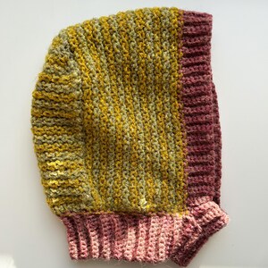 May include: A handmade crochet hood in shades of yellow, green, and mauve. The hood features a ribbed design on the edges and a textured stitch pattern on the main body. The hood is designed to be worn on the head and neck.
