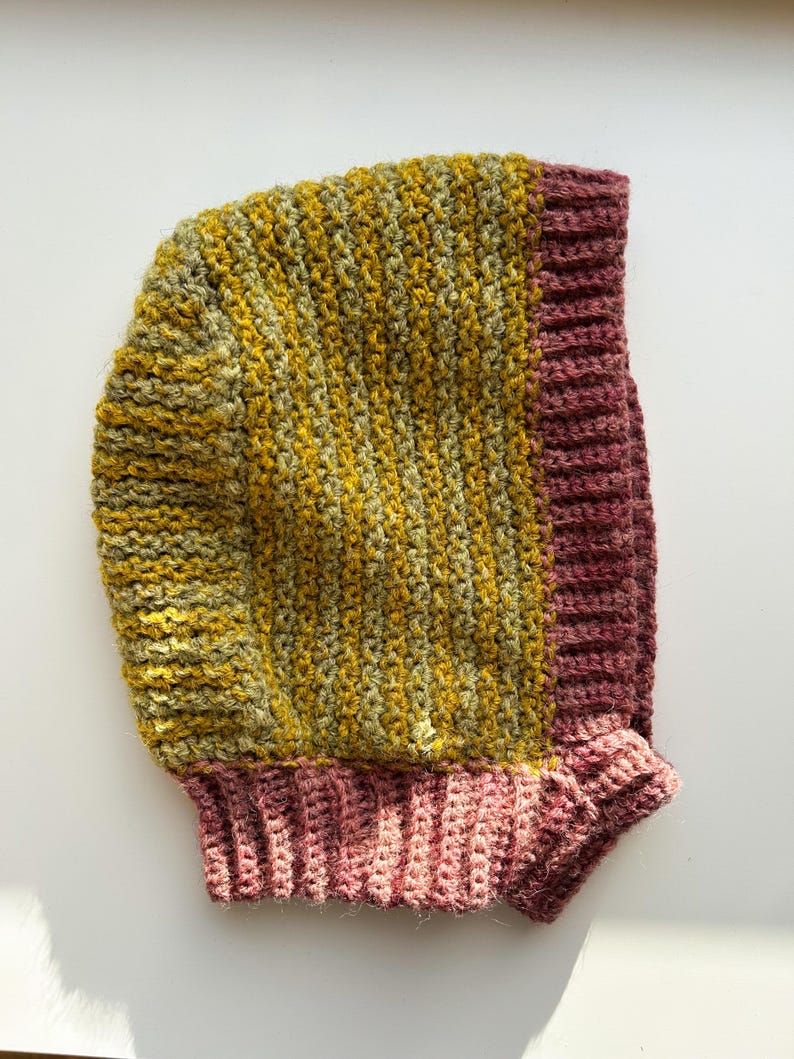 May include: A handmade crochet hood in shades of yellow, green, and mauve. The hood features a ribbed design on the edges and a textured stitch pattern on the main body. The hood is designed to be worn on the head and neck.