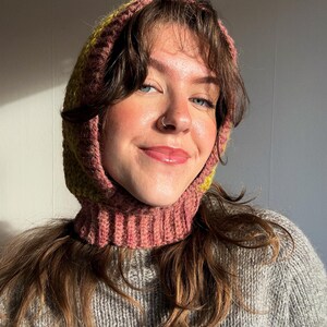 May include: A pink and yellow knitted balaclava with a ribbed neck and hood. The balaclava is worn over a gray sweater. The knitted hood is a cozy accessory for cold weather.