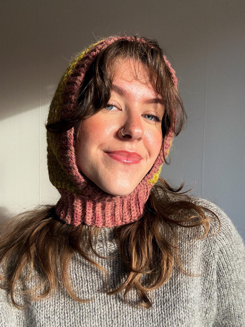 May include: A pink and yellow knitted balaclava with a ribbed neck and hood. The balaclava is worn over a gray sweater. The knitted hood is a cozy accessory for cold weather.