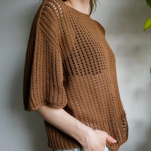 May include: A brown, open-weave knit top with short, puffed sleeves. The top has a round neckline and a loose fit. The knit pattern features vertical and horizontal lines, creating a textured appearance. The top is made of a soft, lightweight material.