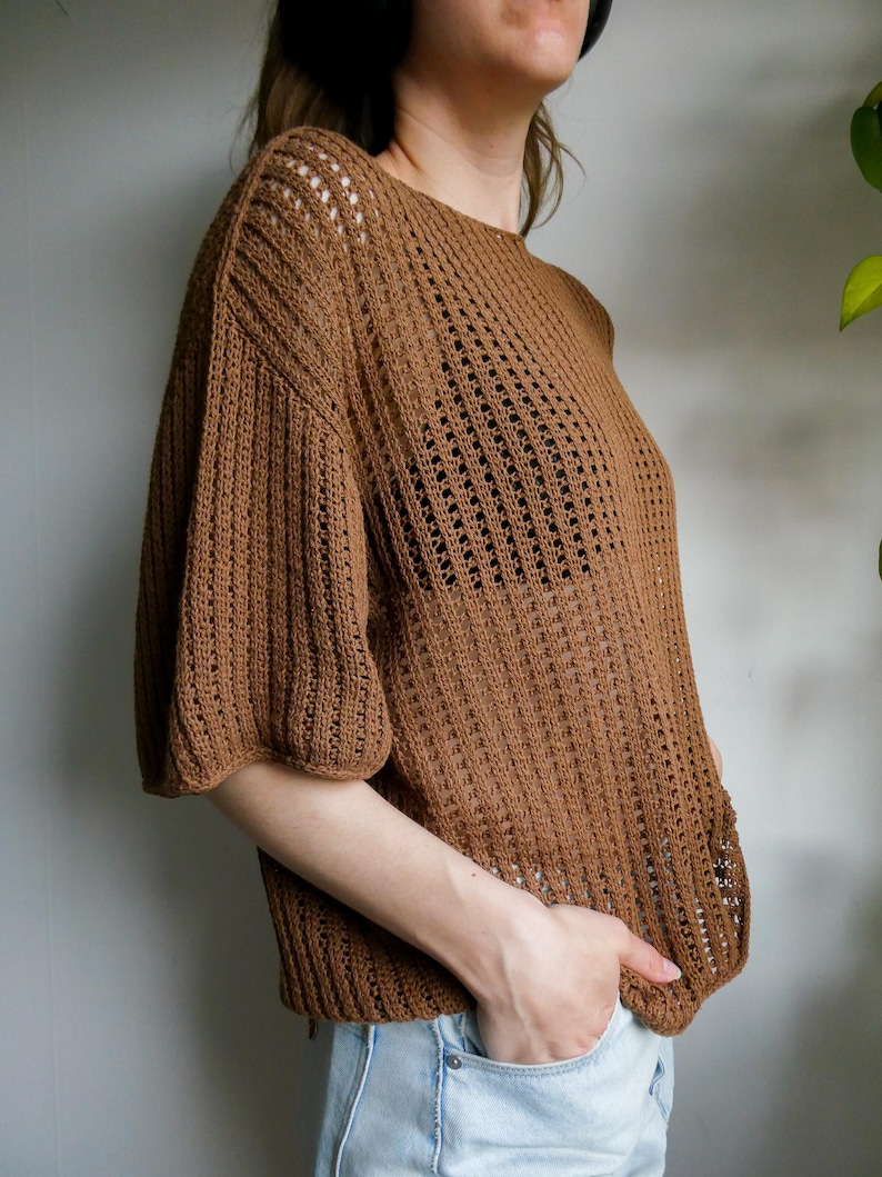 May include: A brown, open-weave knit top with short, puffed sleeves. The top has a round neckline and a loose fit. The knit pattern features vertical and horizontal lines, creating a textured appearance. The top is made of a soft, lightweight material.