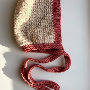 May include: A handmade crochet bonnet in a two-tone design. The bonnet features a light beige center panel and a deep rose-colored border and ties. The textured stitch creates a cozy, handcrafted look. The ties are long and can be tied under the chin.