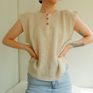 May include: A sleeveless cream-colored knitted sweater with a ribbed texture and pink buttons down the front.