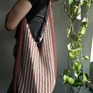 May include: A large, handmade shoulder bag with a brown border and a cream and brown striped pattern. The bag is made of a thick, textured material and has a long strap. The bag is being worn over the shoulder.