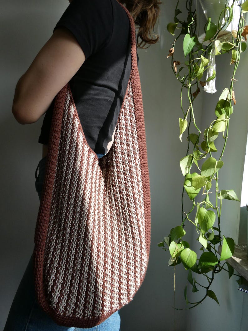 May include: A large, handmade shoulder bag with a brown border and a cream and brown striped pattern. The bag is made of a thick, textured material and has a long strap. The bag is being worn over the shoulder.