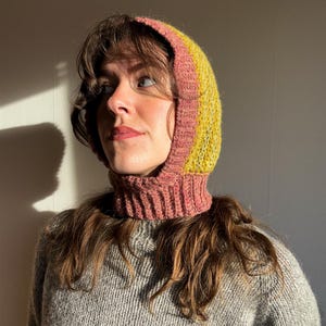 May include: A knitted balaclava in shades of pink, yellow, and green. The hood features a ribbed neck and a striped design. The balaclava is made of a soft, textured yarn, perfect for cold weather. The garment is shown on a person.
