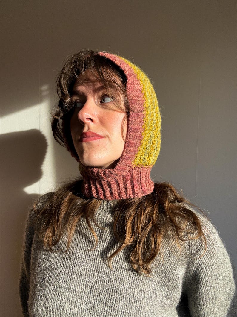 May include: A knitted balaclava in shades of pink, yellow, and green. The hood features a ribbed neck and a striped design. The balaclava is made of a soft, textured yarn, perfect for cold weather. The garment is shown on a person.