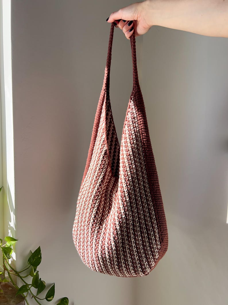 May include: A handmade, open-weave tote bag in a vertical striped pattern of dusty rose and cream. The bag has long, matching straps and a rounded bottom. The bag is held up by a hand.