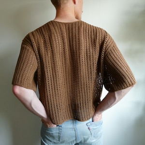 May include: A brown, short-sleeved crochet top with a ribbed design. The top has a relaxed fit and is made of a light brown yarn. The back of the top is visible, showing the texture and construction of the crochet.