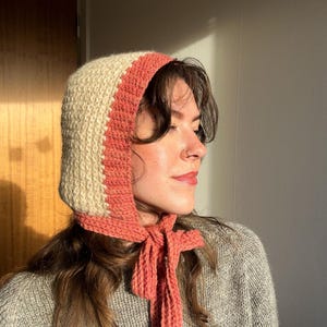 May include: A knitted bonnet in cream and coral. The bonnet has a cream-colored main section with a coral-colored band around the face and a coral tie. The bonnet is made of a soft, textured yarn.