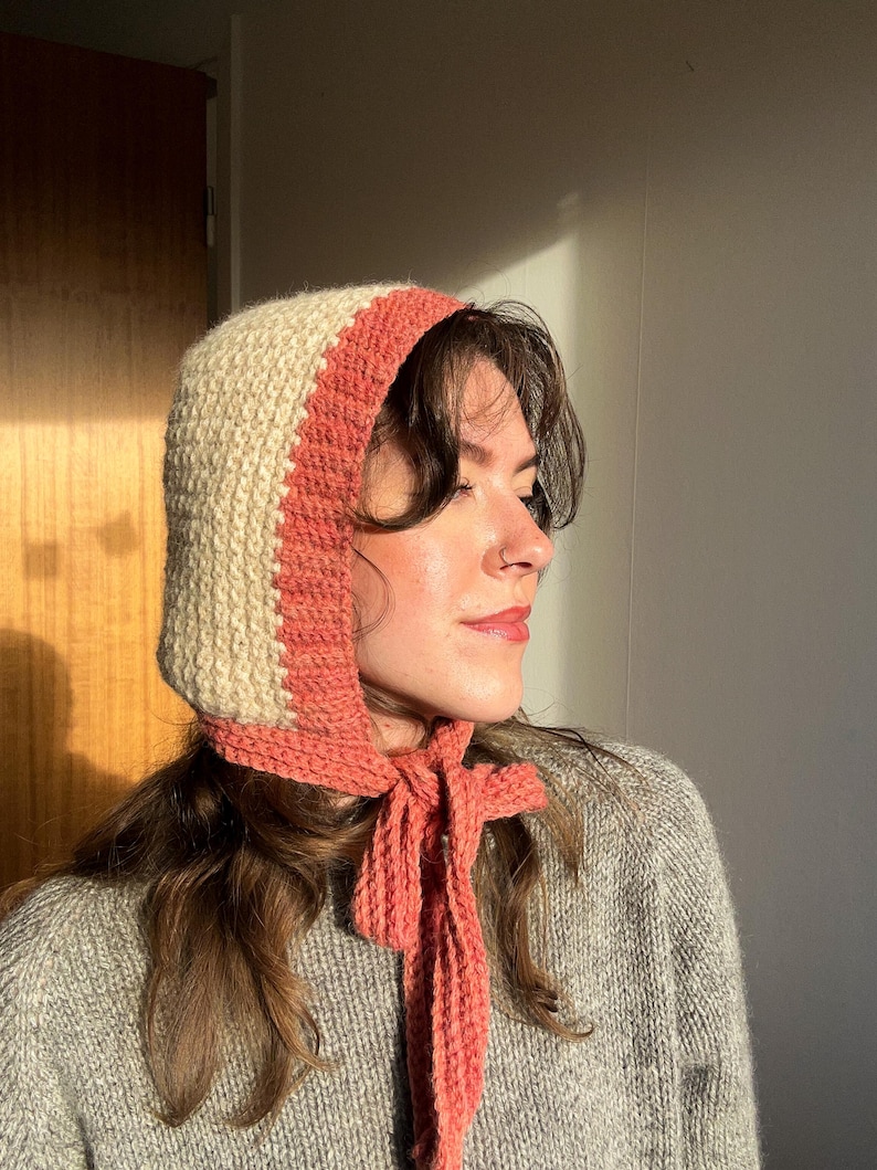 May include: A knitted bonnet in cream and coral. The bonnet has a cream-colored main section with a coral-colored band around the face and a coral tie. The bonnet is made of a soft, textured yarn.