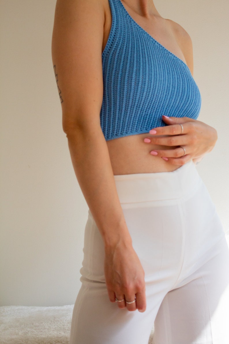 May include: A woman wearing a light blue one-shoulder knitted crop top and white high-waisted trousers.