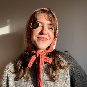 May include: A knitted bonnet in shades of cream, peach, and pink, with long ties that can be tied in a bow. The bonnet is worn over the head and frames the face. The garment is made of a soft, textured yarn.