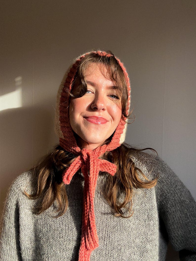 May include: A knitted bonnet in shades of cream, peach, and pink, with long ties that can be tied in a bow. The bonnet is worn over the head and frames the face. The garment is made of a soft, textured yarn.