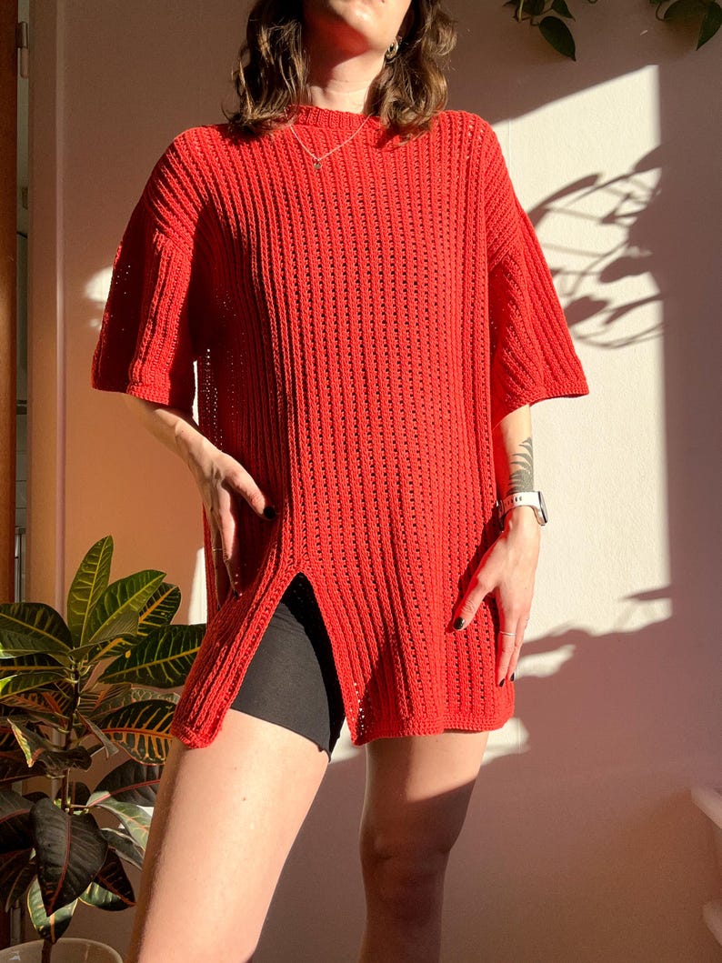 May include: A bright red, short-sleeved knit top with a high side slit. The top has a ribbed texture and a loose fit. The model is wearing black shorts underneath. The top is a vibrant, solid color, perfect for a casual, stylish look.