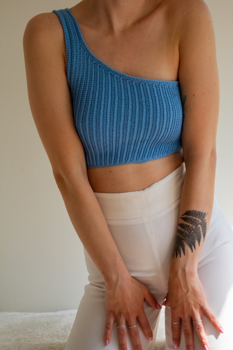 May include: A woman wearing a light blue one-shoulder crop top and white trousers. She has a fern tattoo on her right arm.