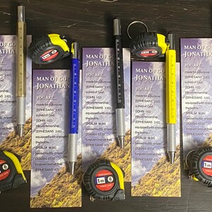 May include: A collection of pens, measuring tapes, and cards with text. The pens have ruler markings and come in silver, blue, black, and yellow. The measuring tapes are black and yellow and measure 1 meter. The cards have the text "MAN OF GOD JONATHAN".