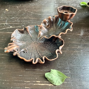 May include: A bronze-toned metal incense holder shaped like a large lotus leaf with a small frog resting on the edge. A smaller, elevated lotus leaf cup sits above the main base, featuring a perforated center for holding incense sticks securely.
