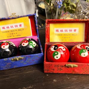 May include: Two sets of Chinese health balls in rectangular fabric-covered boxes. One set is black, the other red, both featuring yin-yang symbols. Each box lid interior displays a yellow card with the text Chinese Health Balls Made in China.