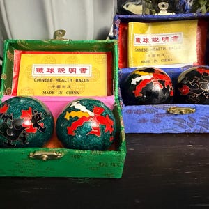 May include: Two sets of Chinese health balls in rectangular fabric-covered boxes. The left box is green with two teal balls featuring black and red dragon designs. The right box is blue with two black balls featuring red and white dragon patterns.