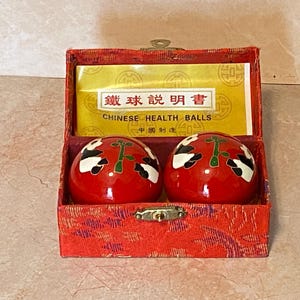 May include: A red box with a gold clasp contains two red Chinese health balls. Each ball has a black and white design with a green accent. The box has a gold label that reads "Chinese Health Balls".