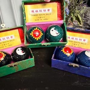 May include: Three sets of Chinese health balls in rectangular boxes. Each set contains two spheres featuring sun and moon designs. The boxes are green, blue, and patterned, each including a yellow instruction card labeled CHINESE HEALTH BALLS.
