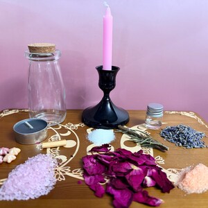 May include: A clear glass bottle with a cork stopper, a black candle holder with a pink taper candle, a small glass vial, a grey tea light, and piles of pink salt, dried rose petals, lavender, white powder, and small star-shaped wax melts.