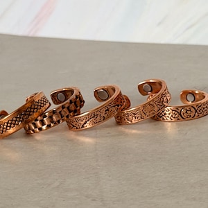 May include: Five copper-colored open-ended rings arranged in a row. Each ring features a distinct embossed pattern, including cross-hatching, geometric squares, and floral motifs. Small circular magnets are visible on the inner ends of each band.