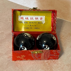May include: A red, rectangular box with a gold clasp, containing two black Chinese health balls. Each ball features a white yin-yang symbol. The box has a gold interior with Chinese text and the words "CHINESE HEALTH BALLS".