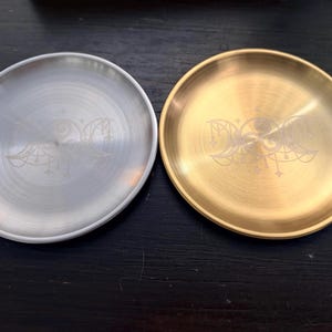 May include: Two circular metal trays, one in a silver tone and one in a gold tone, each featuring a centered laser-engraved triple moon symbol with delicate star accents. The trays have a smooth, brushed metallic finish and raised edges.