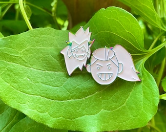 May include: Two enamel pins featuring cartoon characters. The pins are white with black outlines and are set against a green leafy background.
