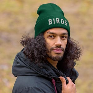 Może przedstawiać: Ciemnozielona czapka z napisem "BIRDS" wyhaftowanym na biało. Czapka wykonana jest z miękkiego, dzianinowego materiału. Osoba na zdjęciu ma na sobie czarną kurtkę.