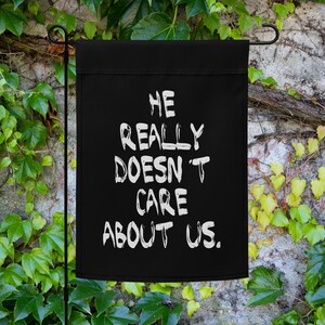 May include: Black garden flag with white text that reads "He really doesn't care about us."