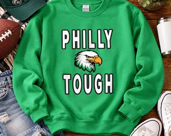 May include: A bright green crewneck sweatshirt featuring the text PHILLY TOUGH in bold white letters with a black outline. Centered between the two words is a graphic illustration of a bald eagle head with white, black, and yellow details.