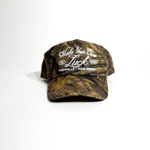May include: Camouflage baseball cap with the text "Make Your Own Luck" in white script. The cap also features the words "Nashville - New Jersey" in a smaller font. The hat is made of a camouflage pattern in shades of brown, green, and tan.