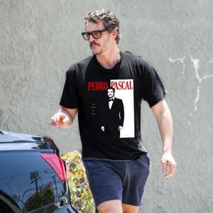 Puede incluir: Un hombre lleva una camiseta negra con un gráfico blanco de Pedro Pascal. El gráfico incluye el texto "Pedro Pascal" en letras rojas y una imagen en blanco y negro de Pedro Pascal con traje. El texto "The  World  Is  Yours" está escrito en letras rojas pequeñas debajo de la imagen.