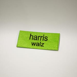 Puede incluir: Un parche rectangular de tela verde lima con texto bordado en negro que dice "harris walz".