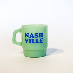 Könnte beinhalten: Eine hellgrüne Keramiktasse mit einer blauen Aufschrift "NASHVILLE".