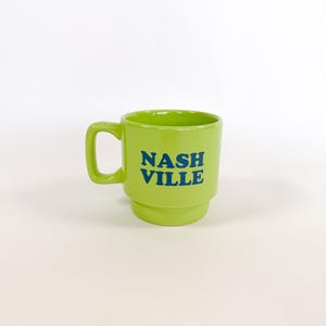 May include: A bright green ceramic mug with the word "NASHVILLE" in blue block letters. The mug has a handle and a stacked design. The mug is a solid, vibrant green color.